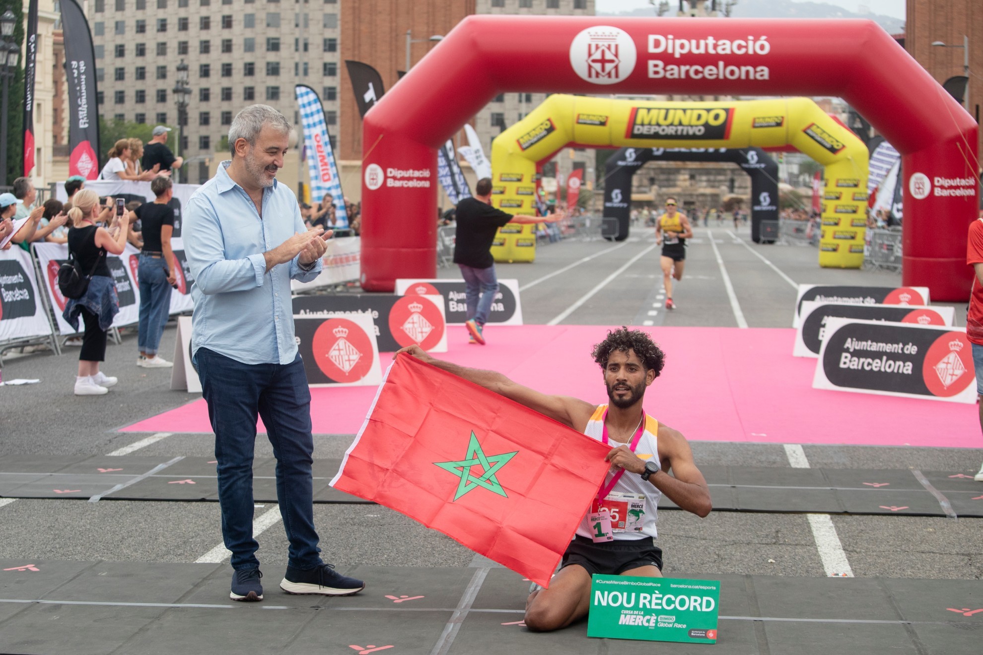 El atleta marroquí Mohamed El Ghazouany cruza la línea de meta como ganador y posa con la bandera del país alahuí junto al alcalde de Barcelona, Jaume Collboni, al término de la Cursa de la Mercè 2023.